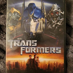 Op de afbeelding: DVD-hoes voor de film Transformers. De hoes toont een close-up van Optimus Prime, Bumblebee en Megatron. De titel "Transformers" is in grote, vetgedrukte letters geschreven. De achtergrond is een stadsbeeld met zonsondergang.