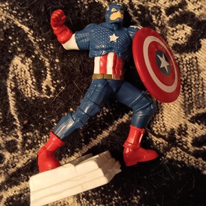 May include: A red, white, and blue action figure of Captain America in a fighting pose. He is holding a red, white, and blue shield in his right hand. The figure is standing on a white base.