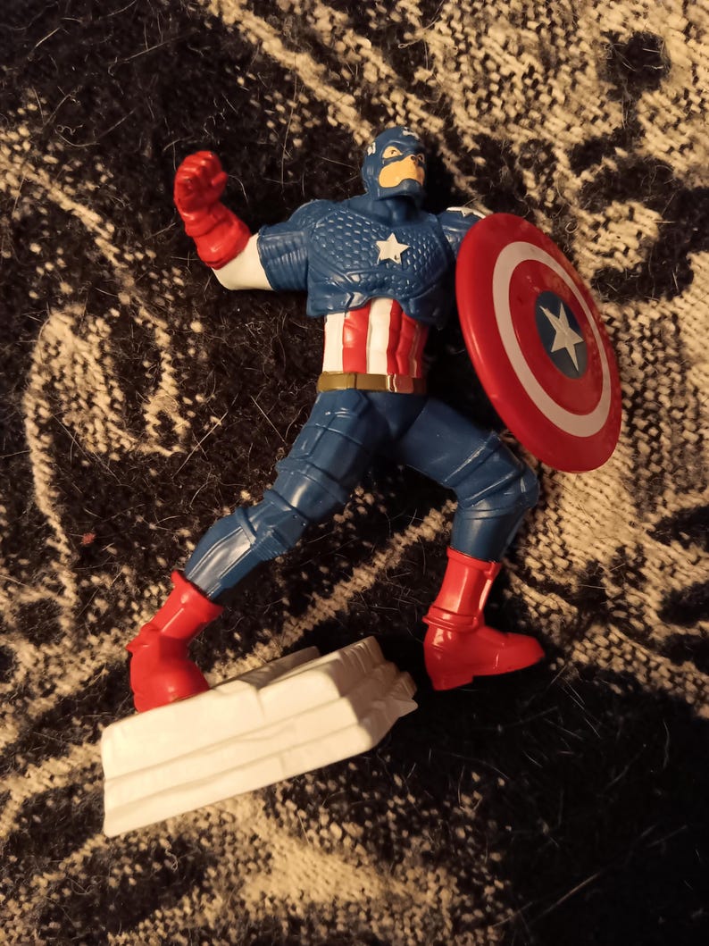 May include: A red, white, and blue action figure of Captain America in a fighting pose. He is holding a red, white, and blue shield in his right hand. The figure is standing on a white base.