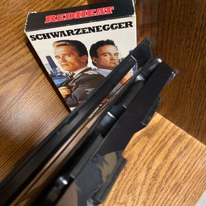 May include: A VHS video cassette tape with a black plastic case. The cover art features a red and white title card that reads "RED HEAT" and "SCHWARZENEGGER". The cover art also features a photo of Arnold Schwarzenegger and James Belushi.