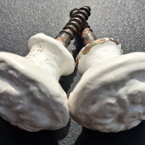 May include: Two white ceramic cabinet knobs with a textured, floral-inspired relief pattern on the front. Each knob features a metal screw stem with visible signs of wear, including dark metallic discoloration and chipped paint on the base.