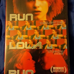 Könnte beinhalten: DVD-Hülle für den Film "Lola rennt". Das Cover hat ein rot-schwarzes Design mit dem Titel in großen silbernen Buchstaben. Das Bild enthält Standbilder aus dem Film und Zitate wie "Dazzling" und "Exhilarating".