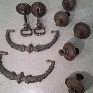 Op de afbeelding: Een assortiment antieke bronzen trekhandgrepen en knoppen. De hardware omvat twee trekhandgrepen met sierlijke handgrepen, twee trekhandgrepen met ronde achterplaten en verschillende knoppen met decoratieve details. De stukken hebben een verweerde, verouderde uitstraling.