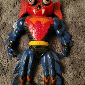 Puede incluir: Una figura de acción de plástico roja y azul con ojos grandes, una placa de pecho negra y un cinturón amarillo. La figura tiene una apariencia de criatura con una cabeza grande y un cuerpo pequeño.
