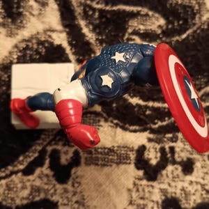 May include: A red, white, and blue plastic action figure of Captain America. The figure is posed with its right arm raised and its left arm bent at the elbow. The figure is holding a red, white, and blue shield in its right hand. The figure is standing on a white base.