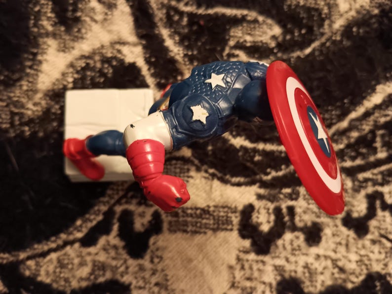 May include: A red, white, and blue plastic action figure of Captain America. The figure is posed with its right arm raised and its left arm bent at the elbow. The figure is holding a red, white, and blue shield in its right hand. The figure is standing on a white base.