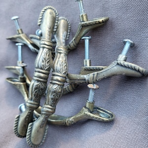 May include: A collection of antique brass-toned furniture pulls. Each pull features a decorative handle with leaf and rope detailing, and a screw for attachment. The pulls are arranged on a gray fabric surface.