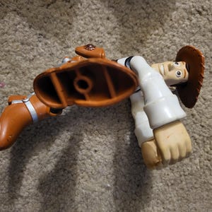 Op de afbeelding: Een plastic speelgoedfiguur van Woody uit de Toy Story-franchise. Woody draagt zijn kenmerkende bruine cowboyhoed, vest en laarzen. Hij staat in een rennende pose met zijn rechterbeen naar voren gestrekt.