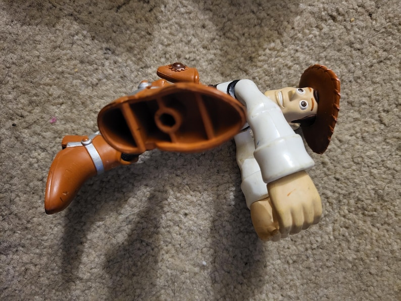 Op de afbeelding: Een plastic speelgoedfiguur van Woody uit de Toy Story-franchise. Woody draagt zijn kenmerkende bruine cowboyhoed, vest en laarzen. Hij staat in een rennende pose met zijn rechterbeen naar voren gestrekt.
