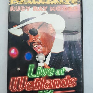 May include: DVD case for "The Dolemite Collection" featuring Rudy Ray Moore. The cover shows Moore in a white hat and sunglasses, holding a microphone. The title "Live at Wetlands N.Y.C." is displayed in neon-style lettering.