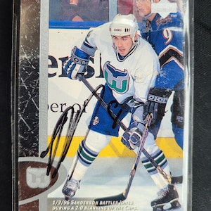 Puede incluir: Tarjeta de hockey con un jugador con camiseta blanca y turquesa. La tarjeta está firmada y tiene el texto "1/3/96 Sanderson Battles Jones During a 2-0 Blanking of the Caps." El jugador sostiene un palo de hockey.