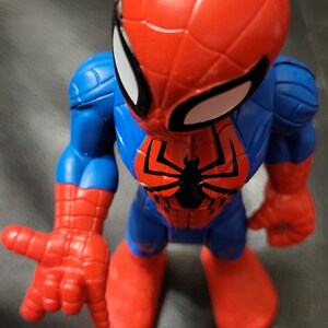 May include: A Spider-Man action figure in a dynamic pose. The figure features a red and blue color scheme, with a large red head and white eye details. The figure's chest has a black spider emblem. The hands and feet are red.