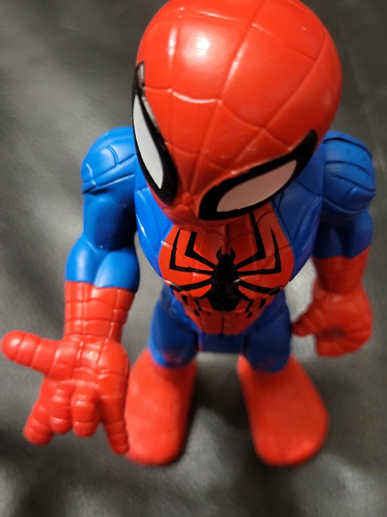 May include: A Spider-Man action figure in a dynamic pose. The figure features a red and blue color scheme, with a large red head and white eye details. The figure's chest has a black spider emblem. The hands and feet are red.