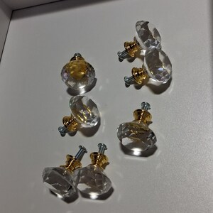 May include: A set of six cabinet knobs featuring clear, faceted, diamond-shaped glass heads attached to polished gold-tone metal bases. Each knob includes a silver-colored metal screw extending from the back for installation purposes.