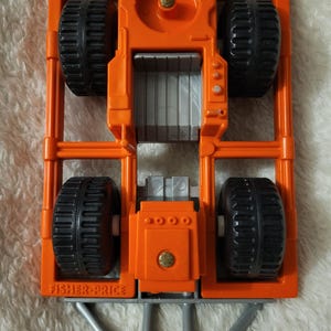 May include: Orange toy truck with black wheels and a gray bed. The truck has a silver metal bumper and the words "Fisher-Price" printed on the side.