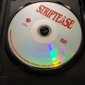 May include: A DVD of the movie "Striptease" in a black case. The disc is white with the title in red, the Warner Bros. logo, and "DVD VIDEO" printed on it. The disc has a reflective surface.