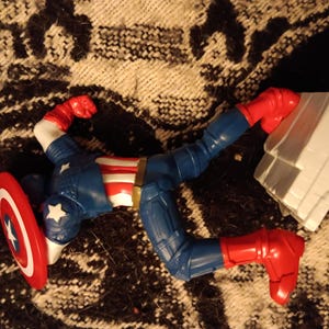May include: A red, white, and blue action figure of Captain America lying on its back with its arms outstretched. The figure is holding a red, white, and blue shield. The figure is lying on a brown and white patterned surface.