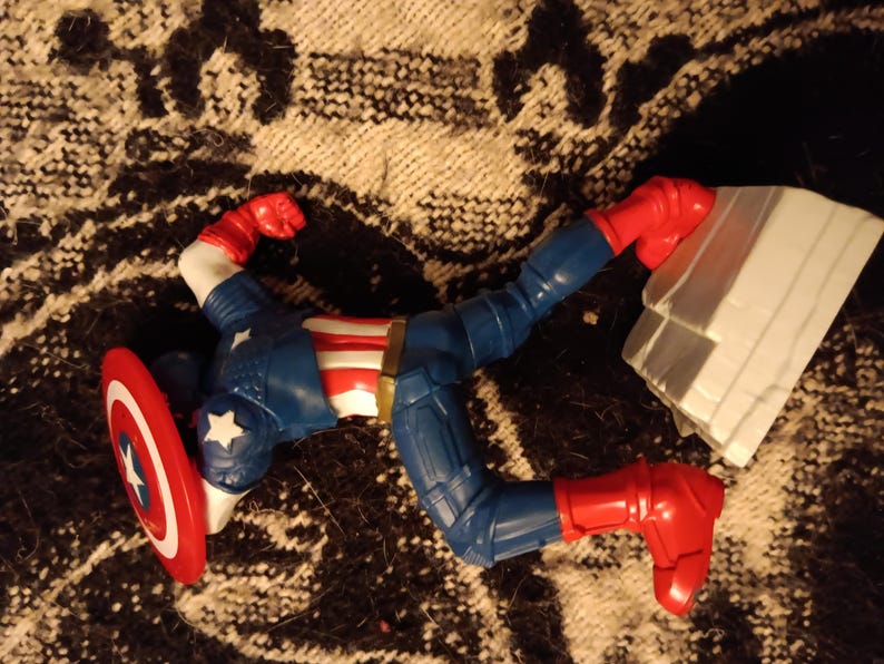 May include: A red, white, and blue action figure of Captain America lying on its back with its arms outstretched. The figure is holding a red, white, and blue shield. The figure is lying on a brown and white patterned surface.