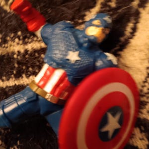 May include: A red, white, and blue action figure of Captain America, wearing a blue and white costume with a red, white, and blue shield. The figure is lying on its side with the shield in front of it.