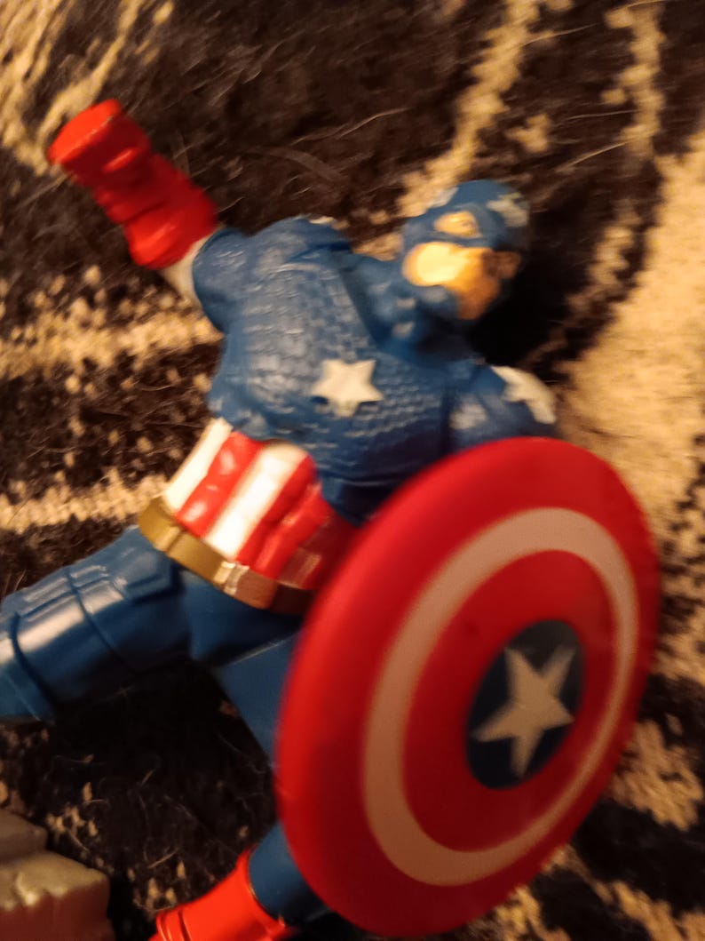 May include: A red, white, and blue action figure of Captain America, wearing a blue and white costume with a red, white, and blue shield. The figure is lying on its side with the shield in front of it.