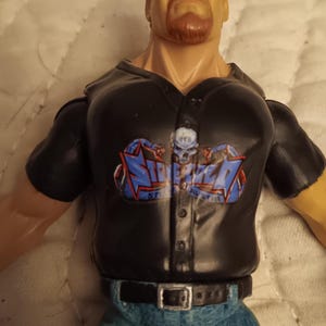 May include: A black action figure wearing a black t-shirt with a blue and white logo that says "Stone Cold Steve Austin" and blue jeans with a black belt.