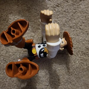 Op de afbeelding: Een plastic speelgoedfiguur van Woody uit de Toy Story-franchise. Woody draagt zijn kenmerkende bruine vest, cowboyhoed en laarzen. Hij bevindt zich in een pose in de lucht met zijn armen omhoog.