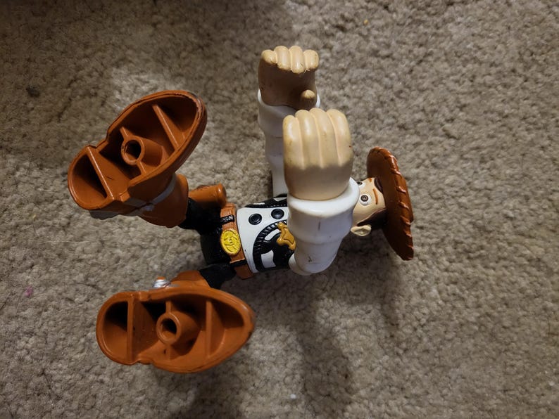 Op de afbeelding: Een plastic speelgoedfiguur van Woody uit de Toy Story-franchise. Woody draagt zijn kenmerkende bruine vest, cowboyhoed en laarzen. Hij bevindt zich in een pose in de lucht met zijn armen omhoog.