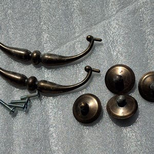 May include: Two curved bronze-toned metal drawer pulls with a smooth finish, four matching circular knobs, and four silver-coloured metal mounting screws. The hardware features a dark, aged metallic patina with subtle variations in colour tone.