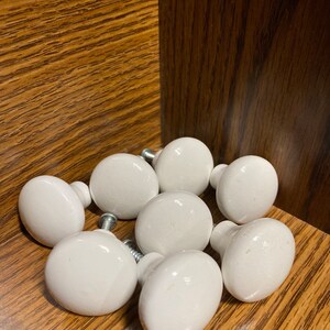 May include: Eight white ceramic cabinet knobs with silver screws. The knobs are round and have a smooth finish. They are arranged in a circle on a wooden surface.
