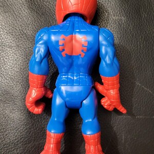 May include: A blue and red Spider-Man action figure. The figure has a red helmet, red gloves, and red boots. The back of the figure has a red spider emblem. The figure is posed with its arms at its sides.