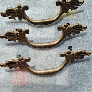 May include: Three antique bronze drawer pulls with ornate floral and leaf designs. Each pull has a curved handle and is attached with screws. The pulls have a weathered appearance, suggesting age and use.