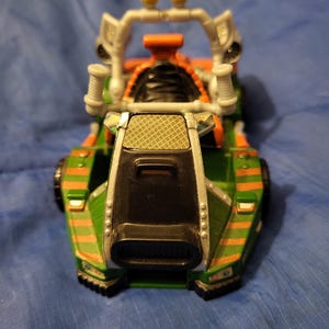 Può includere: Un veicolo giocattolo verde e arancione con una griglia anteriore nera e un roll-bar argentato. Il giocattolo ha due luci gialle sulla parte superiore e strisce arancioni. Il veicolo è su una superficie blu.