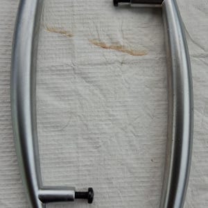 May include: Two curved, silver-toned metal cabinet handles with a smooth, matte finish. Each handle features two cylindrical mounting points with black threaded bolts extending outward, designed for attachment to furniture or cabinetry doors.
