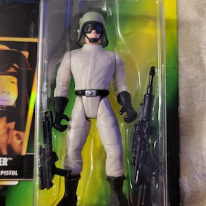 Può includere: Action figure di Star Wars di un personaggio in uniforme grigio chiaro, guanti e stivali neri, con due fucili neri. La figura è in una confezione di plastica trasparente con le parole "The Power of the Force" visibili.