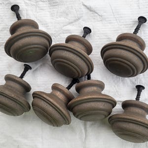 Puede incluir: Un conjunto de siete pomos redondos para armarios con un acabado texturizado en tonos bronce oscuro. Cada pomo presenta un diseño circular escalonado y está unido a un tornillo de metal negro para su montaje. Los pomos tienen un aspecto desgastado.