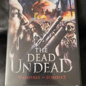 Puede incluir: Caja de DVD de "The Dead Undead: Vampires vs Zombies". La portada presenta un primer plano de la cara de un vampiro, una horda de zombis y un paisaje urbano en llamas. El título está en letras blancas grandes.