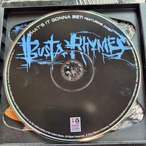 Può includere: Un CD nero con le parole "WHAT'S IT GONNA BE?!" e "BUSTA RHYMES" in blu. Il CD è in un astuccio nero. Il testo "FEATURING JANET" è anch'esso visibile.