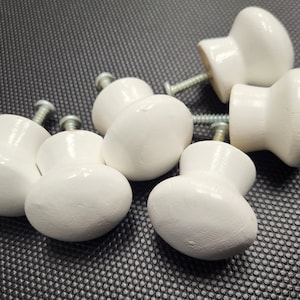 Pode incluir: Um conjunto de seis puxadores brancos de madeira para armários, com parafusos prateados. Os puxadores, de formato arredondado e em forma de cogumelo, estão dispostos sobre uma superfície texturizada cinza escuro. Ideal para restauração de móveis.