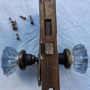 May include: A vintage mortise door lock set featuring two clear, fluted glass knobs with aged brass-toned metal bases and a matching rectangular brass faceplate with a latch mechanism, accompanied by five small, separate brass-colored screws.