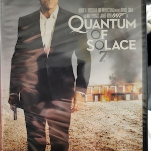 May include: DVD case for the James Bond film "Quantum of Solace." The cover features a man in a black suit holding a gun, with the film's title in large white letters. The background shows a fiery scene.