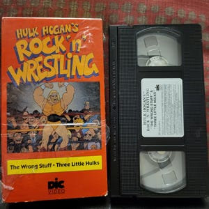 May include: A VHS tape and a black VHS cassette. The tape box is orange with the title "Hulk Hogan's Rock 'n' Wrestling" and cartoon illustrations. The tape itself has a white label with the title and episode names.