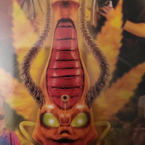May include: A red and orange alien-like creature with green eyes and a long, thin body. The creature has a large, round head with a small mouth and a pointed nose. The creature's body is covered in scales and has two long, thin arms. The creature is standing in front of a bright, yellow background.