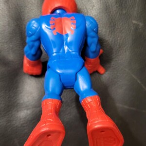 May include: A blue and red action figure of a superhero, viewed from the back. The figure has a red helmet, red boots, and red gloves. A red spider emblem is on the back. The figure is lying down on a dark surface.