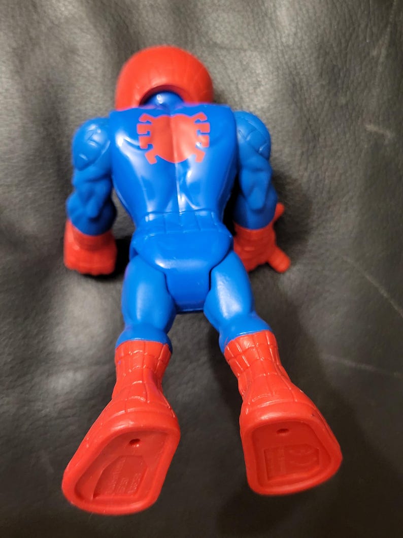May include: A blue and red action figure of a superhero, viewed from the back. The figure has a red helmet, red boots, and red gloves. A red spider emblem is on the back. The figure is lying down on a dark surface.