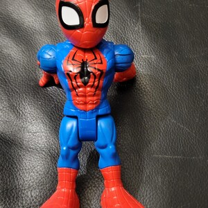 May include: A Spider-Man action figure in a standing pose. The figure is primarily red and blue, with a black spider emblem on the chest. The head is red with white eye shapes.