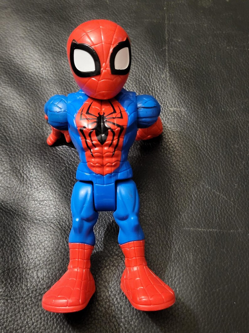 May include: A Spider-Man action figure in a standing pose. The figure is primarily red and blue, with a black spider emblem on the chest. The head is red with white eye shapes.