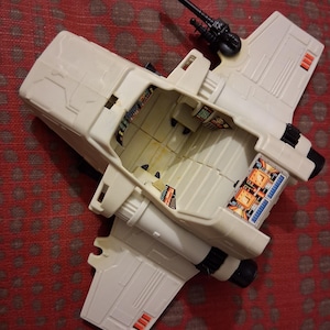 Könnte beinhalten: Ein cremefarbenes Spielzeugraumschiff im Vintage-Stil mit schwarzen und orangefarbenen Akzenten. Das Spielzeug hat einen detaillierten Innenraum mit farbenfrohen Paneelen und eine schwarze Kanone oben. Die Flügel haben rote und schwarze Details. Ein klassisches Spielzeug für Sammler.