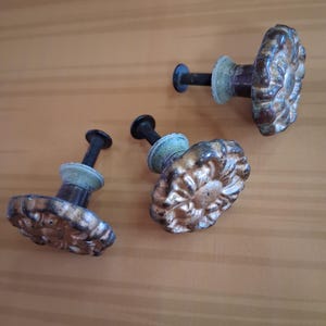 May include: Three antique bronze cabinet knobs with a floral design. The knobs have a distressed finish with hints of green and are mounted on black screws. The knobs are arranged on a light brown wooden surface.