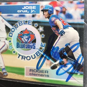 Puede incluir: Tarjeta de béisbol con José Cruz Jr. con el uniforme de los Blue Jays de Toronto. La tarjeta tiene el texto "DOUBLE TROUBLE" y "ROGER CLEMENS". El fondo es un estadio con otros jugadores.
