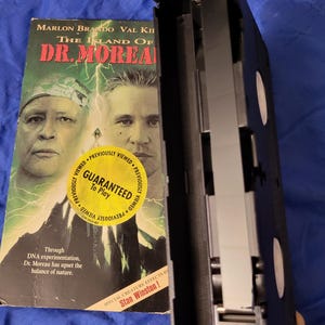 Puede incluir: Una cinta VHS de "La Isla del Dr. Moreau" con Marlon Brando y Val Kilmer. La portada muestra el título de la película y las caras de los actores, con un adhesivo "Garantizado para reproducir". La cinta negra está abierta, mostrando la cinta magnética.
